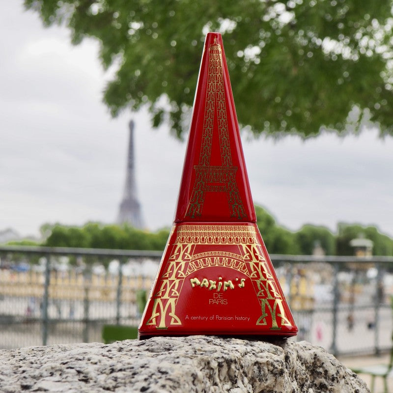 Red Eiffel Tower tin with gold detailing on a stone surface, blurred background of trees and buildings.